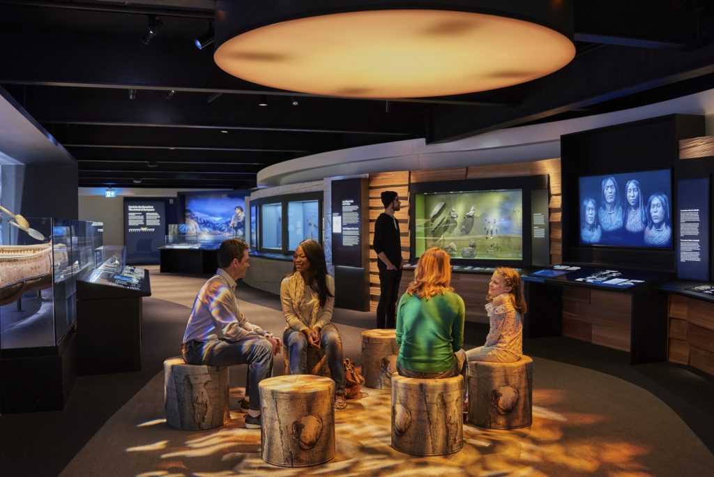 Shishalh Nation exhibition module in the Canadian History Hall. Four people are seated on imitation stumps in the gallery space with digital interactives around them.